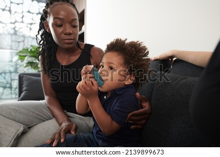 Worried mother looking at son using inhaler when having asthma attack at home Royalty-Free Stock Photo #2139899673