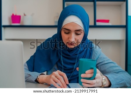 A Muslim business woman working on a laptop in the home desk. Muslim girl student in hijab having online education. Confident Muslim woman drinks coffee while having video call conference.	