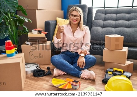 Young hispanic woman moving to a new home sitting on the floor doing happy thumbs up gesture with hand. approving expression looking at the camera showing success. 