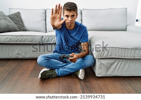 Young handsome hispanic man playing video game sitting by the sofa with open hand doing stop sign with serious and confident expression, defense gesture 