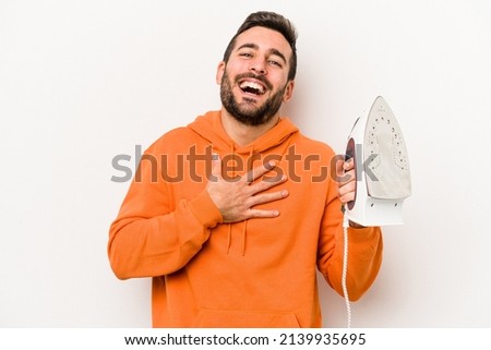 Young caucasian man holding an iron isolated on white background laughs out loudly keeping hand on chest.