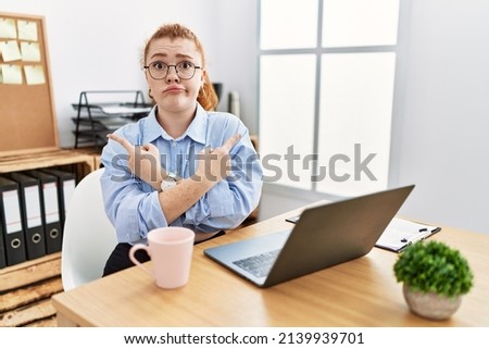 Young redhead woman working at the office using computer laptop pointing to both sides with fingers, different direction disagree 
