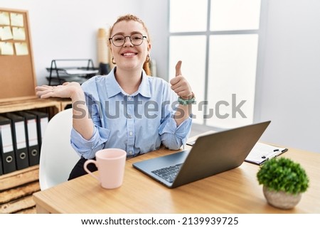 Young redhead woman working at the office using computer laptop showing palm hand and doing ok gesture with thumbs up, smiling happy and cheerful 