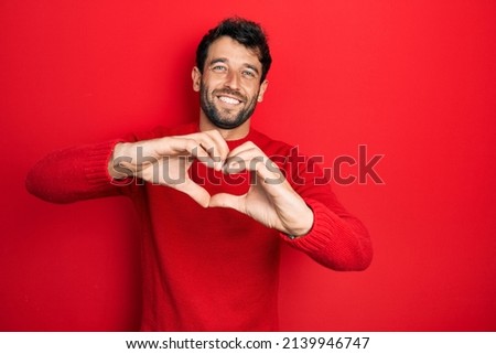 Handsome man with beard wearing casual red sweater smiling in love doing heart symbol shape with hands. romantic concept. 