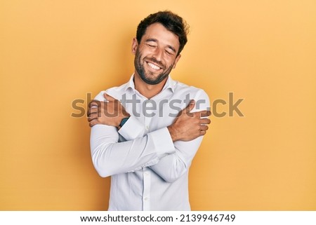 Handsome man with beard wearing casual white t shirt hugging oneself happy and positive, smiling confident. self love and self care  Royalty-Free Stock Photo #2139946749