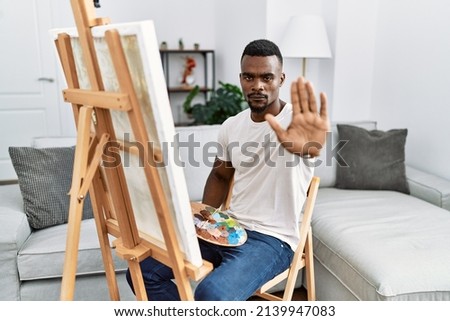 Young african man painting on canvas at home doing stop sing with palm of the hand. warning expression with negative and serious gesture on the face. 