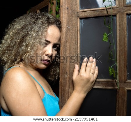 Brunette woman wearing a blue dress, in a frosted glass window and wooden frame looking towards the camera in an early evening with light from left to right. Mention of female empowerment.