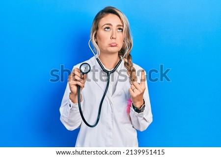 Beautiful young blonde doctor woman holding stethoscope pointing up looking sad and upset, indicating direction with fingers, unhappy and depressed. 