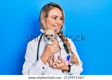 Young blonde veterinarian woman wearing uniform and stethoscope holding small chihuhua with love
