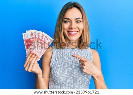 Young caucasian blonde woman holding icelandic krona banknotes smiling happy pointing with hand and finger  Royalty-Free Stock Photo #2139961721