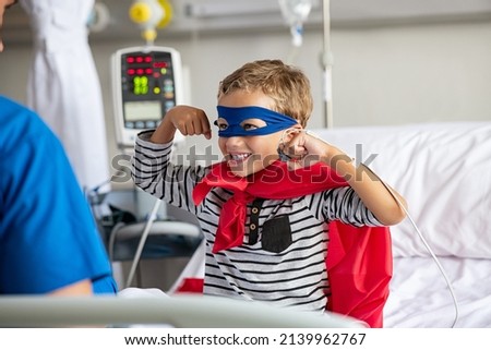 Cheerful strong little boy wearing blue eyeband and red cape like superhero playing with nurse. Playful child gesturing dressed in superhero costume at clinic overcome adversity and health challenge.  Royalty-Free Stock Photo #2139962767