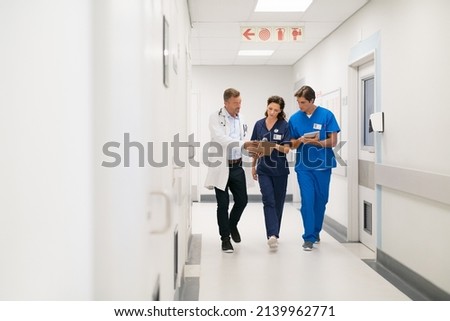 Medical team walking in hurry and interacting at clinic. Mature doctor and surgeon working on digital tablet with nurse and walking in hospital hallway. Head physician working with his medical team. Royalty-Free Stock Photo #2139962771