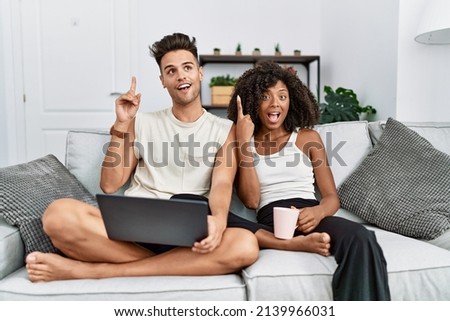 Young interracial couple using laptop at home sitting on the sofa pointing finger up with successful idea. exited and happy. number one. 