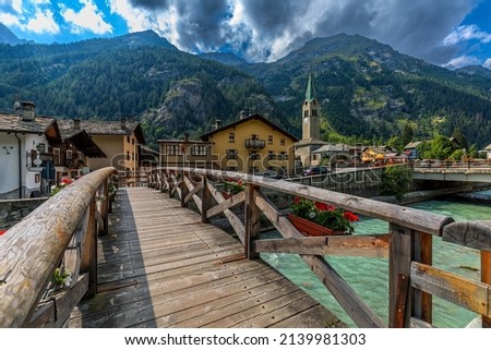 Wooden bridge over mountain river as houses and old church on background in small town of Gressoney-Saint-Jean in Aosta Valley, Italy. Royalty-Free Stock Photo #2139981303