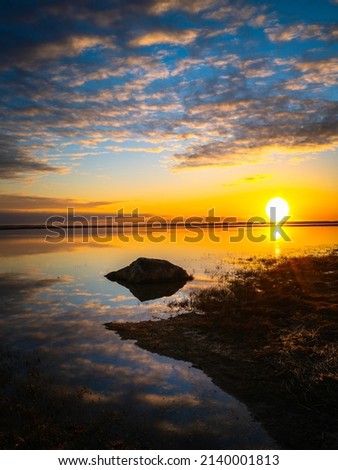 Sunrise, cumulous clouds, beach grass, black rock, reflections in the water, white sun, orange horizon, and blue sky. Tranquil Zen-like seascape on Cape Cod.