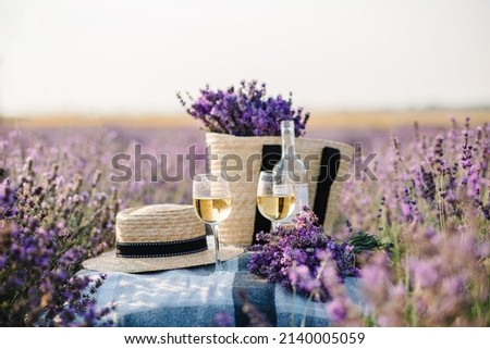 Two glasses with white wine and bottle on background of a lavender field. Straw hat and basket with flowers lavender on a blanket on picnic. Romantic evening in sunset rays. Summer in Provence, France Royalty-Free Stock Photo #2140005059