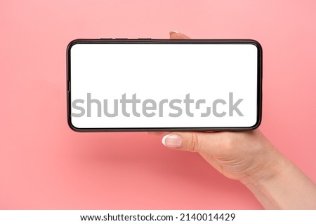 Hand holding mobile phone in horizontal position for mockup. watching streaming video on cellphone on pink table background. Close up of horizontal black smartphone with blank screen in woman hands Royalty-Free Stock Photo #2140014429