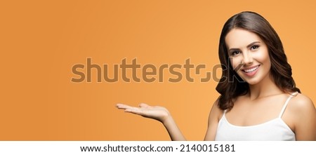 Photo of smiling beautiful woman in white casual wear holding hand with flat palm, giving, advertising some product showing mock up copy space free area. Vivid brown color background. Brunette girl.