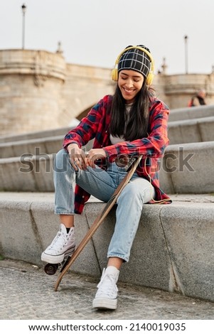 young woman sitting with skateboard. smiling latina listening to music.