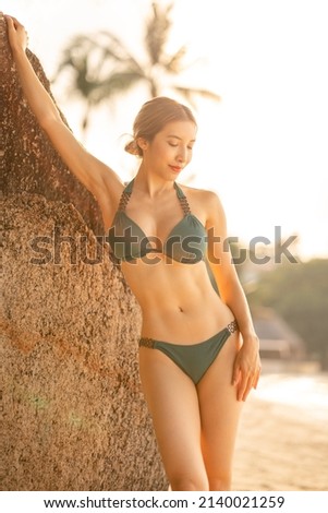Woman in green bikini enjoying a sunset on the beach while standing near rock by the ocean.