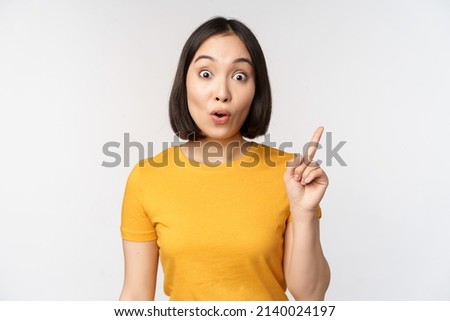 Beautiful young asian woman pointing finger up, smiling and looking amused at camera, showing advertisement, announcement on top, white background