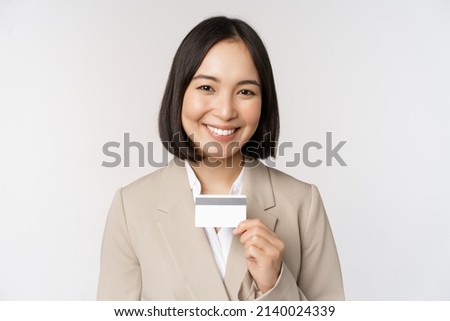 Smiling office clerk, asian corporate woman showing credit card, standing over white background in beige suit