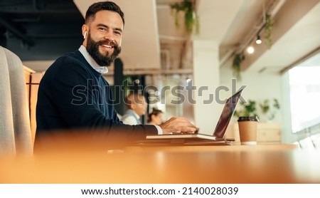 Businessman working on his laptop in a co-working space. Modern businessman smiling at the camera while typing on his laptop and listening to music. Happy entrepreneur sitting in an office lobby. Royalty-Free Stock Photo #2140028039