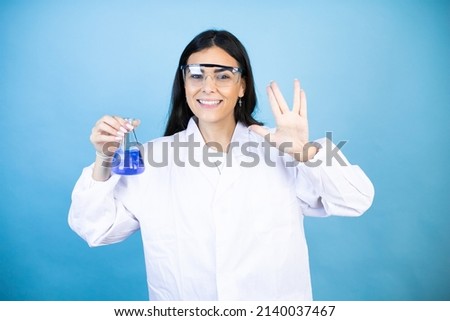Young brunette woman wearing scientist uniform holding test tube over isolated blue background doing hand symbol