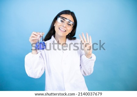 Young brunette woman wearing scientist uniform holding test tube over isolated blue background showing and pointing up with fingers number four while smiling confident and happy