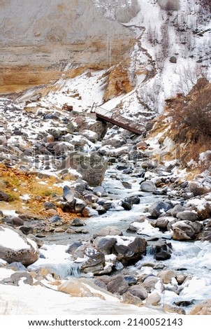 A mountain river with a bridge on the background of a snow-covered slope and a steep rock wall.