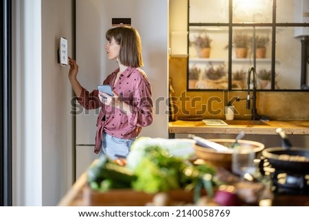 Woman using digital tablet hanging on the wall in the kitchen, controlling smart home devices. Modern kitchen interior with food in front