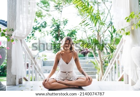 Young smiling woman practicing yoga and meditating in resort hotel outdoor. Concept of tourism, vacation and weekend. Beautiful caucasian girl. Idyllic and tranquil lifestyle on Bali island. Sunny day