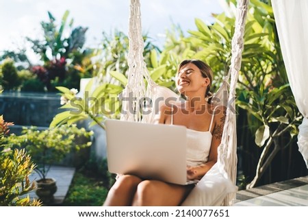 Young joyful woman with laptop rest and enjoy sunny day on hammock chair on terrace of resort hotel. Tourism, vacation and weekend. Pretty caucasian girl with tattoos. Idyllic and tranquil lifestyle Royalty-Free Stock Photo #2140077651