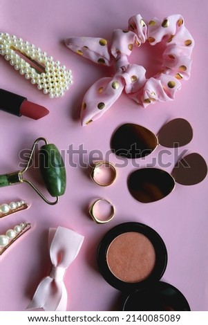 Pink scrunchie, gold earrings and rings, blush, various hair barrettes, lipstick and jade roller on bright pink background. Flat lay. 