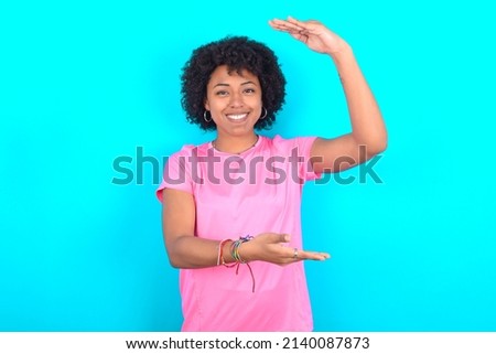 young girl with afro hair style wearing sport pink t-shirt over blue background gesturing with hands showing big and large size sign, measure symbol. Smiling looking at the camera.
