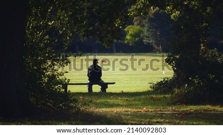 Man sat alone on park bench in silhouette, surrounded by trees and foliage Royalty-Free Stock Photo #2140092803