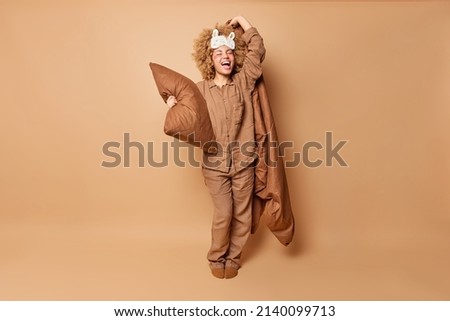 Indoor shot of happy positive young woman stands in full length wears pajama sleepmask holds pillow and blanket has fun after healthy sleep isolated over brown background. Sleeping time concept Royalty-Free Stock Photo #2140099713