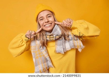 Glad young woman points at toothy smile has white even teeth wears hat sweatshirt and scarf around neck feels very happy isolated over yellow studio background. Human facial expressions concept