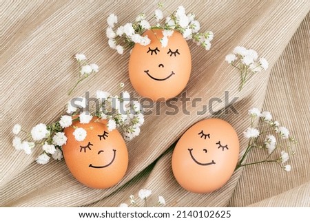 Three happy Easter eggs with cute faces in floral wreath crowns on brown burlap background. Easter eggs with flowers and sleepy eyes in sunny light. Happy Easter concept.