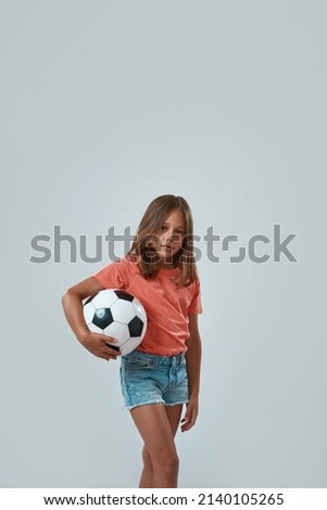 Serious little girl standing with football ball and looking at camera. Caucasian female child wearing pink t-shirt and jean shorts. Childhood concept. White background. Studio shoot. Copy space