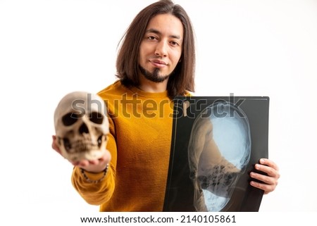 Long-haired bearded caucasian man holding artificial skull and skull x-ray while looking to the camera. Studio shot over white background. High quality photo