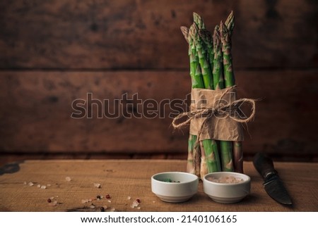 rustic picture with a fresh bunch of asparagus tied with rope on top of a wooden chopping board with salt, pepper and a knife next to it