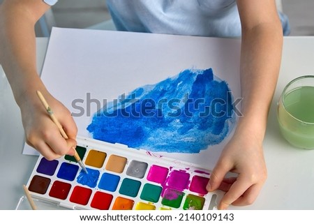 Close up of a child 's hands drawing with a brush and blue paint at a white table. Top view of a boy painting with watercolor paint. Development of creative potential in young children. 