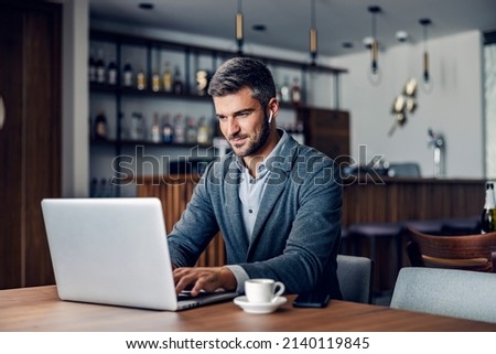 A dedicated busy man dressed in smart casual is sitting in working friendly cafe and typing a report on the laptop. He has earphones in his ears. A businessman typing on a laptop in a cafe. Royalty-Free Stock Photo #2140119845