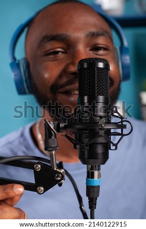 Closeup of professional microphone in front of talking vlogger wearing wireless headphones recording audio podcast. Close focus on studio mic used by influencer to record social media content.
