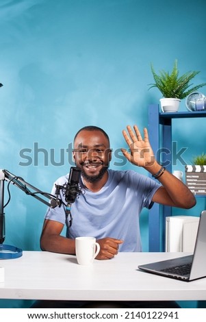 Portrait of african american vlogger waving hand at camera in recording studio with professional microphone and broadcasting setup. Content creator saying hello at audience during online live show.