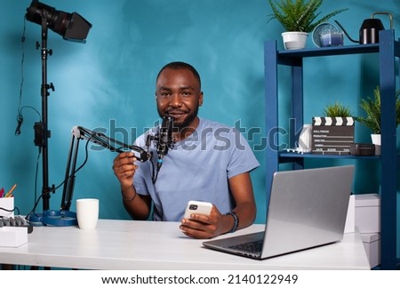 Vlogger holding smartphone looking at camera with hand on microphone sitting at podcast recording desk. Influencer talking into professional mic after reading messages from fans on mobile.