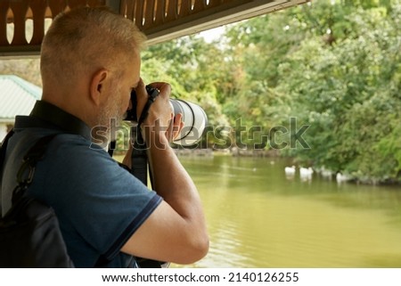 An adult photographer takes pictures with a camera with a long-focus lens of a flock of wild ducks on a pond.                               