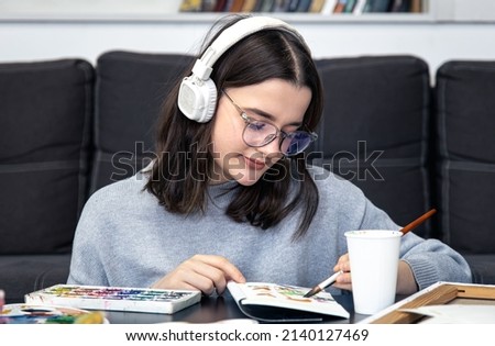 A stylish young woman artist in glasses and headphones draws with paints at home.
