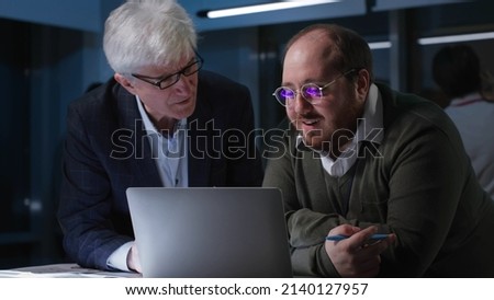 Executive and employee discuss project on laptop in office working late. Business colleagues brainstorm using computer